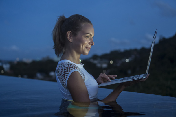 Szabadban álló szép blonde boldog nő laptop számítógép a tetőtéri feszített víztükrű medence felett homályos hegy és az ég háttere az este folyamán. Szabadúszó és technológia - Fotó, kép