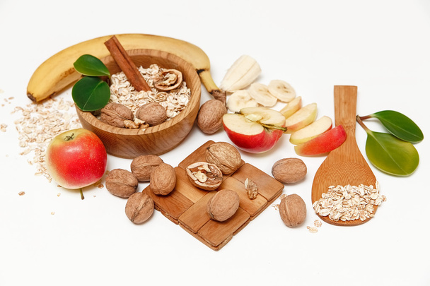 There are Banana,Apple, Walnuts in the Wooden Plate and Rolled Oats,Wooden Spoon,Trivet,with Green Leaves,Healthy Fresh Organic Food on the White Background - Photo, Image