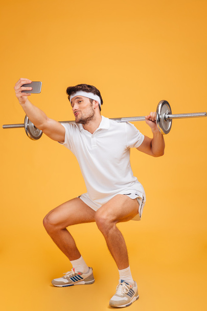 Funny sportsman exercising with barbell and talking selfie using smartphone - Фото, изображение