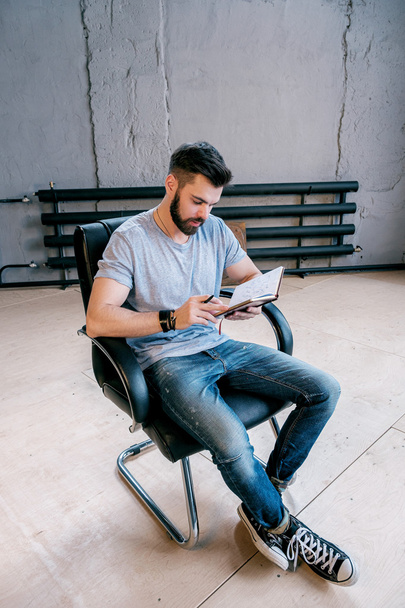 Ritratto di giovane uomo d'affari che analizza il pianificatore quotidiano e siede su una sedia in pelle al coperto
 - Foto, immagini