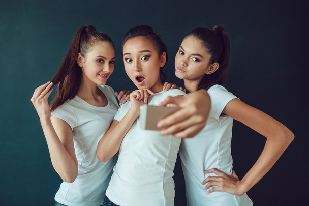 Positive friends portrait of happy girls making selfie, sure funny faces, grimaces, joy, emotions, casual style, pastel colors. Dark background. - Foto, Imagen