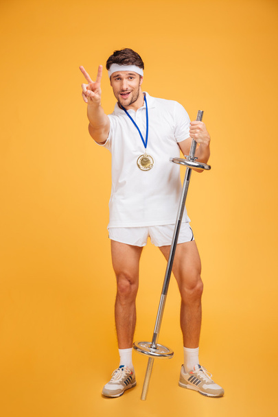 Happy young sportsman with barbell standing and showing victory sign - Zdjęcie, obraz