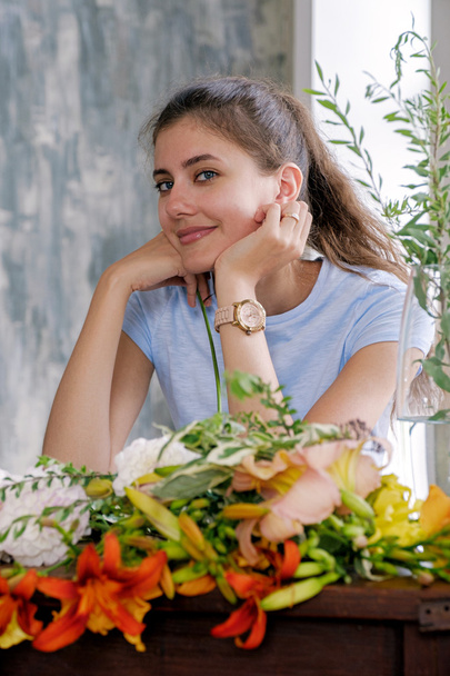 Portrait of pretty young girl with brunette hair in tail smiling at camera at table with bright flowers - Foto, immagini