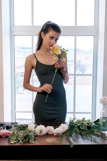 Portrait of young florist with hair in tail smelling beautiful flower while standing at table with fresh cut flowers against of window - Photo, image