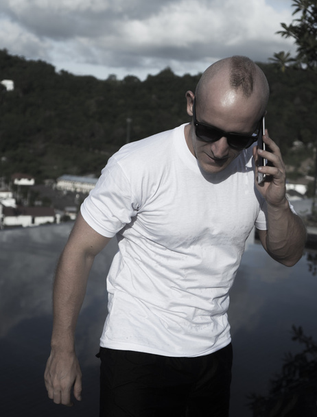 Retrato al aire libre de un joven guapo con gafas de sol hablando por teléfono móvil mientras está de pie en una piscina infinita en la azotea sobre el cielo azul y el paisaje de la ciudad. Independiente y tecnología
 - Foto, imagen