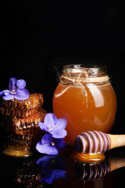 Мед в стеклянной банке на черном фоне. Рядом с фиолетовыми цветами и сотовыми сотами. Натуральный продукт пчеловодства
. - Фото, изображение