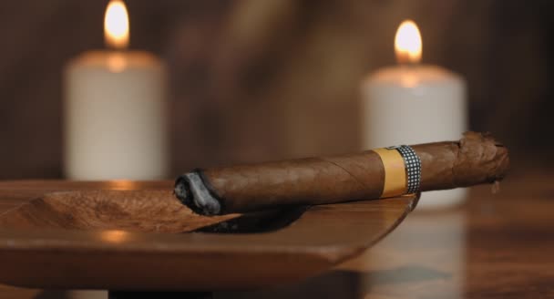 4k: beweging van de schuifregelaar van een gloeiende Cubaanse sigaar in een asbak met sommige kaarsen op de achtergrond naar een open doos sigaren op rustieke houten tafel.  - Video