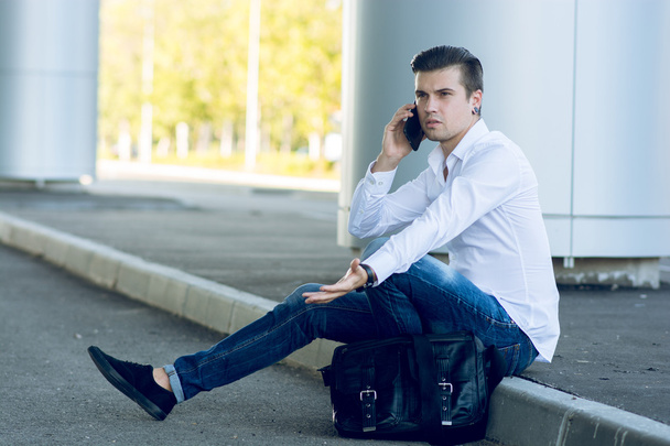 joven guapo hablando teléfono inteligente mientras está sentado cerca de la carretera
 - Foto, imagen