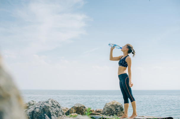 athletic girl drinks water from a bottle on the shore after a workout - Foto, Bild
