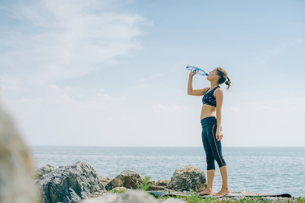 athletic girl drinks water from a bottle on the shore after a workout - Photo, Image