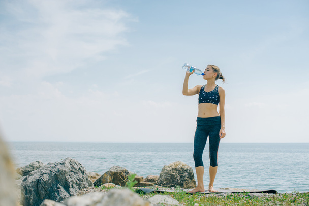спортивна дівчина п'є воду з пляшки на березі після тренування
 - Фото, зображення