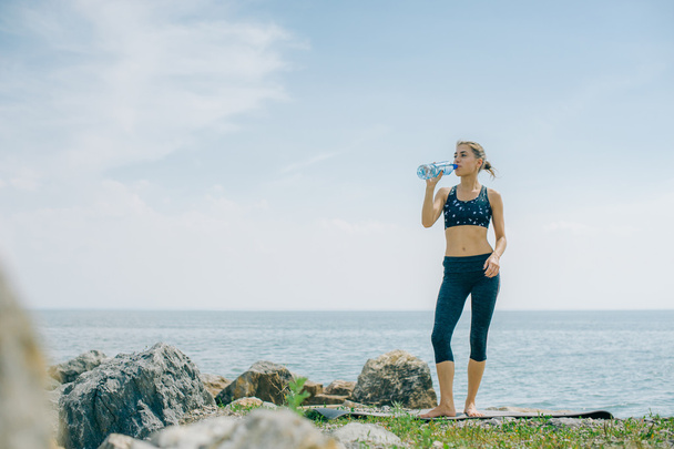 athletic girl drinks water from a bottle on the shore after a workout - Foto, afbeelding