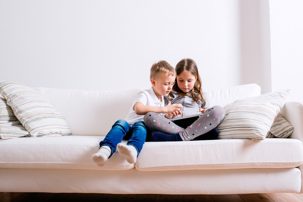 Children at home sitting on sofa, playing with tablet - Фото, изображение