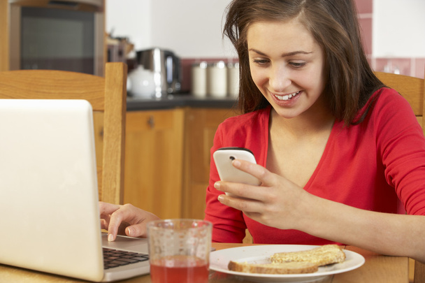 Teenage Girl Using Laptop And Mobile Phone Whilst Eating Breakfa - Foto, Bild