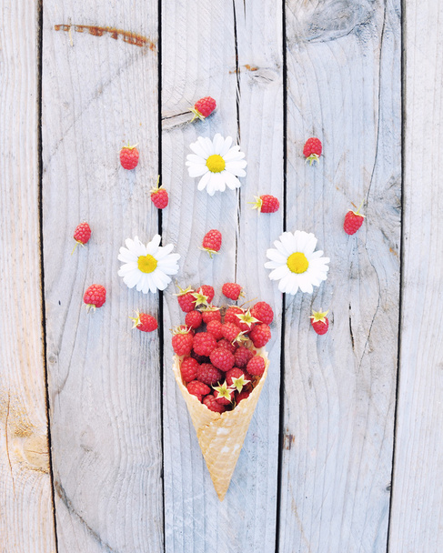 Ripe fresh raspberries and white chamomile flowers in ice cream cone on rustic wooden background. Stylish flat lay. Minimal concept. - Photo, image