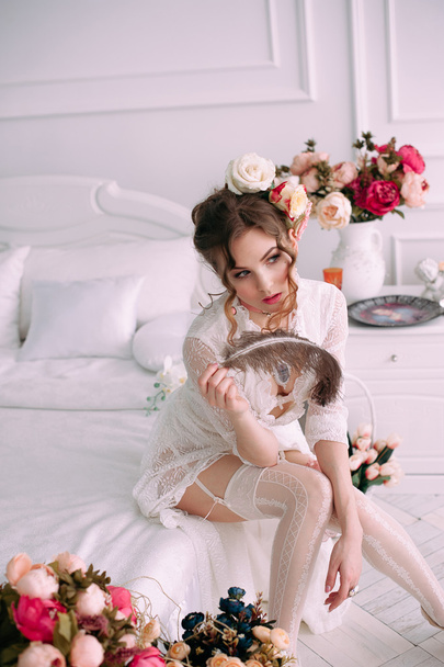 Красива молода сексуальна жінка сидить на білому ліжку, одягнена в біле мереживне плаття, волосся прикрашене квітами. Ідеальний макіяж. Красива мода. Вії. Студійний ретушований знімок
.  - Фото, зображення