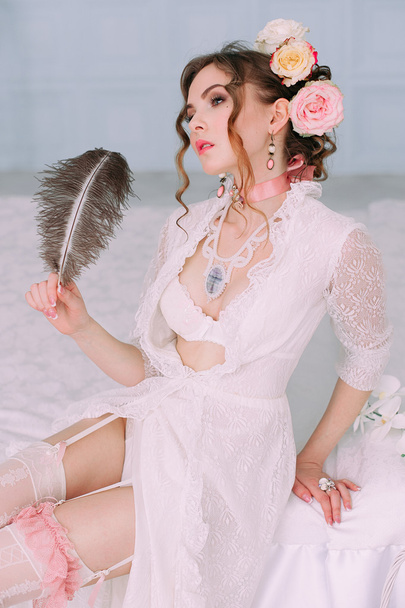 Belle jeune femme sexy assise sur un lit blanc, portant une robe en dentelle blanche, des cheveux décorés de fleurs. Maquillage parfait. Mode beauté. Des cils. Studio retouché
.  - Photo, image