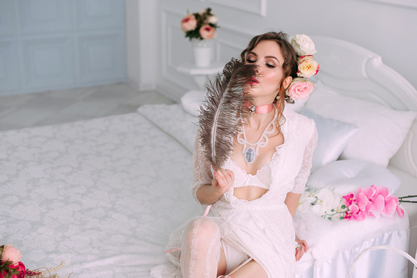 Krásná mladá sexy žena sedící na bílé posteli, na sobě bílé krajkové šaty, vlasy, květinovou výzdobou. Perfektní make-up. Móda krása. Řasy. Studio retušované shot.  - Fotografie, Obrázek