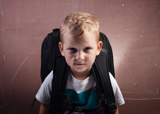 Kleiner Junge mit einem großen schwarzen Rucksack, ein Typ mit blonden Haaren posiert an einer Tafel, - Foto, Bild