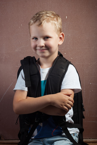Μικρό αγόρι με ένα μεγάλο μαύρο σακίδιο, ένας άντρας με ξανθά μαλλιά ποζάρει στο πίνακα κιμωλίας, - Φωτογραφία, εικόνα