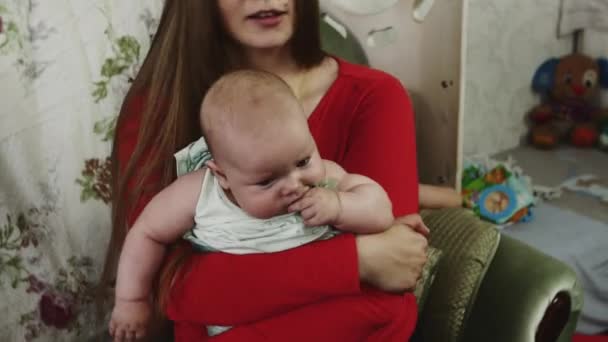 Kırmızı gömlekli anne bebek koltukta oturan ellerini tutun. Annelik. Küçük oğlu. - Video, Çekim