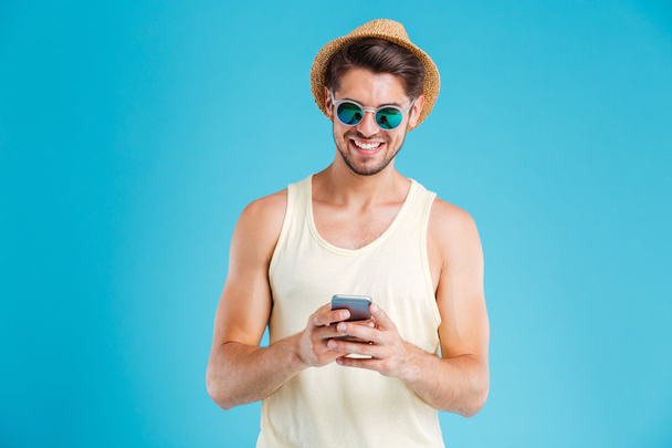 Happy young man in hat and sunglasses using smartphone - Foto, afbeelding