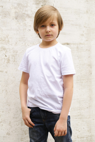 χαριτωμένο ξανθό αγόρι με τζιν και ένα άσπρο πουκάμισο θέτοντας σε φόντο τοίχο - Φωτογραφία, εικόνα