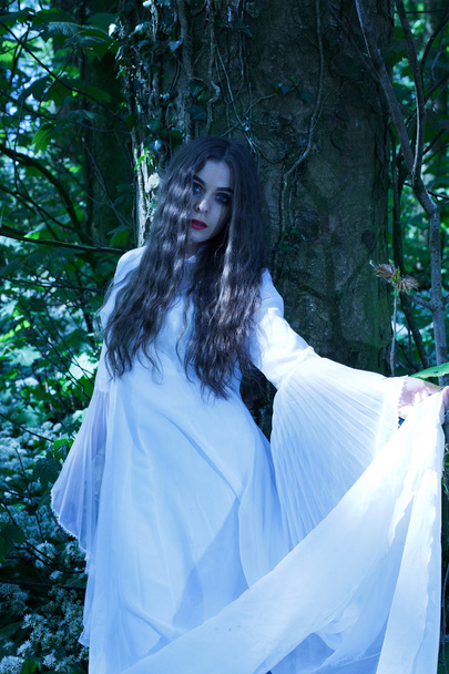 Beautiful woman wearing a long white dress standing against a tree - Photo, Image