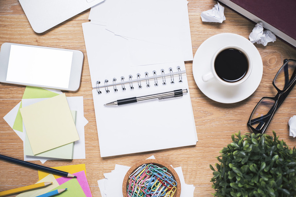Messy workplace with blank white smartphone, open spiral notepad, coffee cup, glasses, plant and various stationery items. Mock up - Photo, image