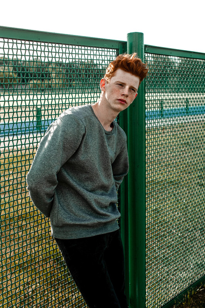 Un giovane bel ragazzo dai capelli rossi alla moda in piedi vicino allo stadio in giornata di sole all'aperto, indossando abbigliamento casual
 - Foto, immagini