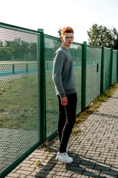 One young handsome red haired fashionable boy standing near stadium in sunny day outdoor , wearing casual clothing - Fotó, kép