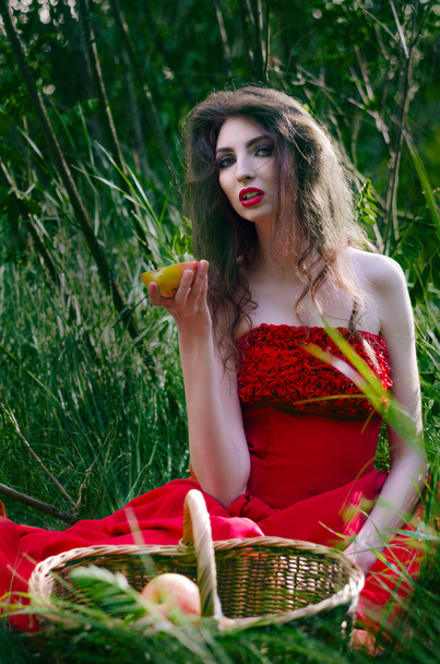 Belle fille, sorcière assise sur l'herbe dans une robe rouge et manger la moitié d'une pomme qui prend au panier
 - Photo, image