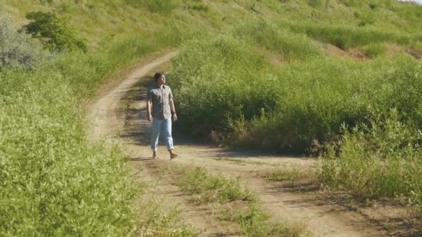 Young handsome barefeet man walking on sand trail between grassy hills at sunny day - Footage, Video
