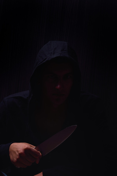 Closeup portrait of a young man in a hoodie, holding a knife in  - Foto, Bild