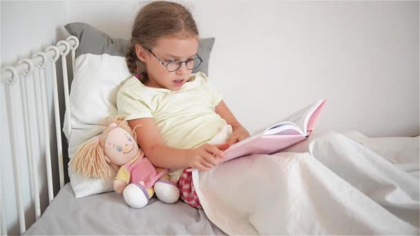 Little girl with glasses leafing through a book while lying in bed. Perhaps a child is sick. Next to her sits a doll. - Кадры, видео