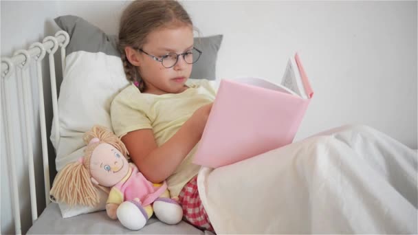Little girl in glasses reading a book while lying in bed. Next to her sits a doll. - Video