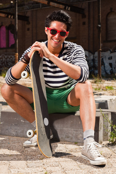 Urban asian man with red sunglasses and skateboard sitting on street. Good looking. Cool guy. Wearing blue white striped sweater and green shorts. Old neglected building in the background. - Foto, imagen