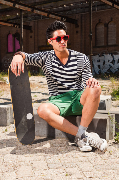 Urbain asiatique homme avec des lunettes de soleil rouges et skateboard assis sur la rue. Beau look. Cool mec. Portant un pull rayé bleu blanc et un short vert. Ancien bâtiment négligé en arrière-plan
. - Photo, image
