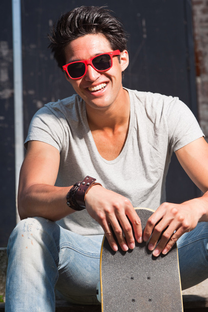 Urban uomo asiatico con occhiali da sole rossi e skateboard seduto sulle scale. Bell'aspetto. Che figo. Indossare camicia grigia e jeans
. - Foto, immagini