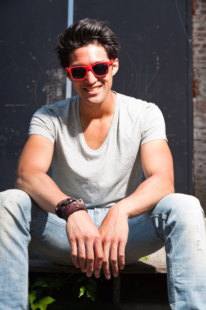 Urban asian man with red sunglasses sitting on stairs. Good looking. Cool guy. Wearing grey shirt and jeans. Old neglected building in the background. - Фото, зображення
