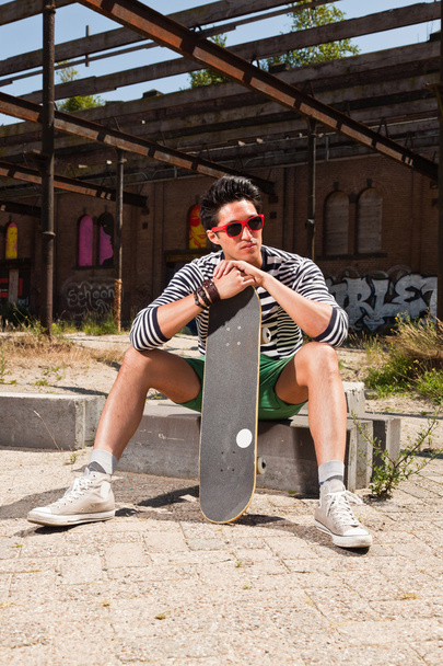 Urban asian man with red sunglasses and skateboard sitting on street. Good looking. Cool guy. Wearing blue white striped sweater and green shorts. Old neglected building in the background. - Фото, зображення