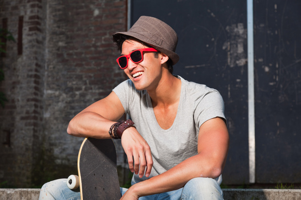 Ein urbaner asiatischer Mann mit Hut, roter Sonnenbrille und Skateboard sitzt auf einer Treppe. gut aussehend. Cooler Typ. trägt graues Hemd und Jeans. altes, vernachlässigtes Gebäude im Hintergrund. - Foto, Bild