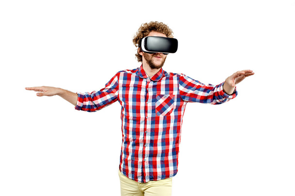 Curly-haired jongeman in geruite hemd met behulp van een Vr headset en het ervaren van virtuele realiteit geïsoleerd op witte achtergrond - Foto, afbeelding