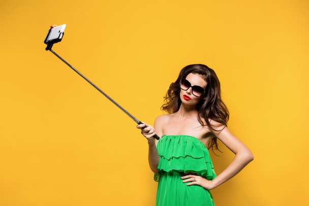 Portrait of young model in green dress and sunglasses posing while making selfie against yellow background. - Fotografie, Obrázek