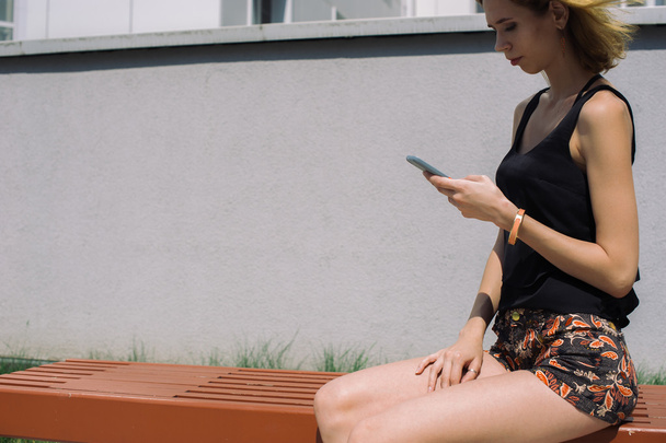 Young slim girl in a black T-shirt using smartphone sitting in the yard - Fotó, kép