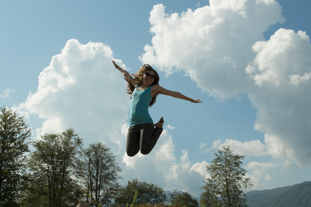 Красивая темноволосая счастливая молодая женщина, прыгающая высоко в воздухе, на фоне летнего голубого неба. горизонтальная картина
 - Фото, изображение