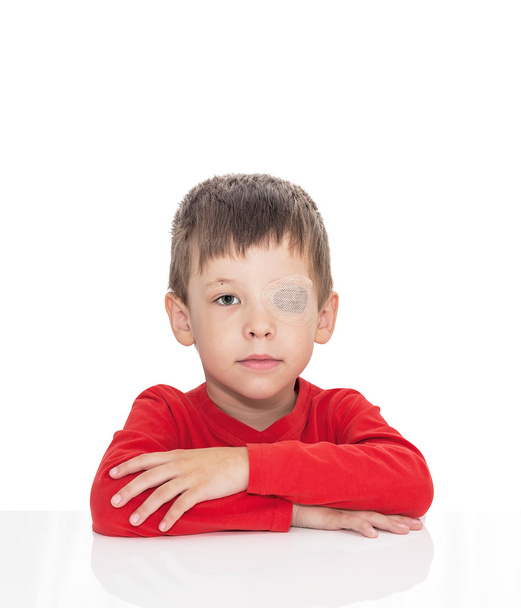 The five-year-old boy sits at a white table, the left eye is stuck with a plaster - Fotografie, Obrázek
