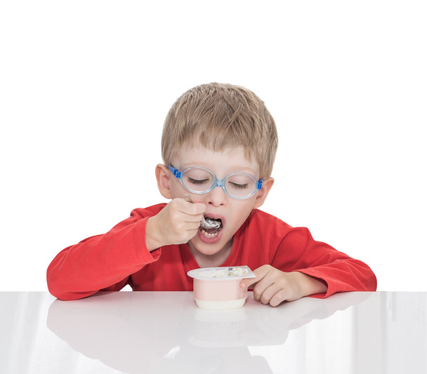 The five-year-old boy sits at a white table and eats yogurt - Fotografie, Obrázek
