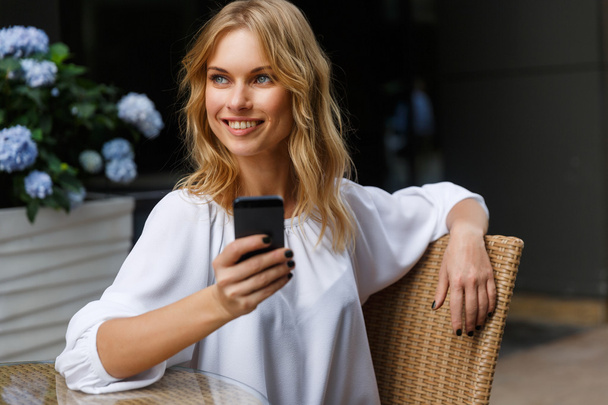 Smiling blond woman in white blouse with phone in hand - Photo, Image