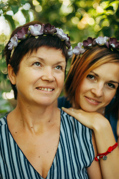 笑顔の母と娘の肖像画の木の背景に屋外から造花の花輪でポーズをとっています。家族の概念。母と娘の関係. - 写真・画像